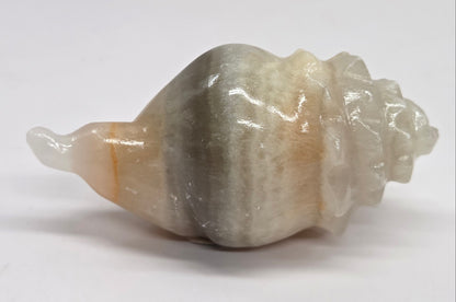 A carved stone shell sculpture with a spiral seashell shape, featuring a smooth, polished finish in shades of gray, beige, and white, set against a plain light gray background.