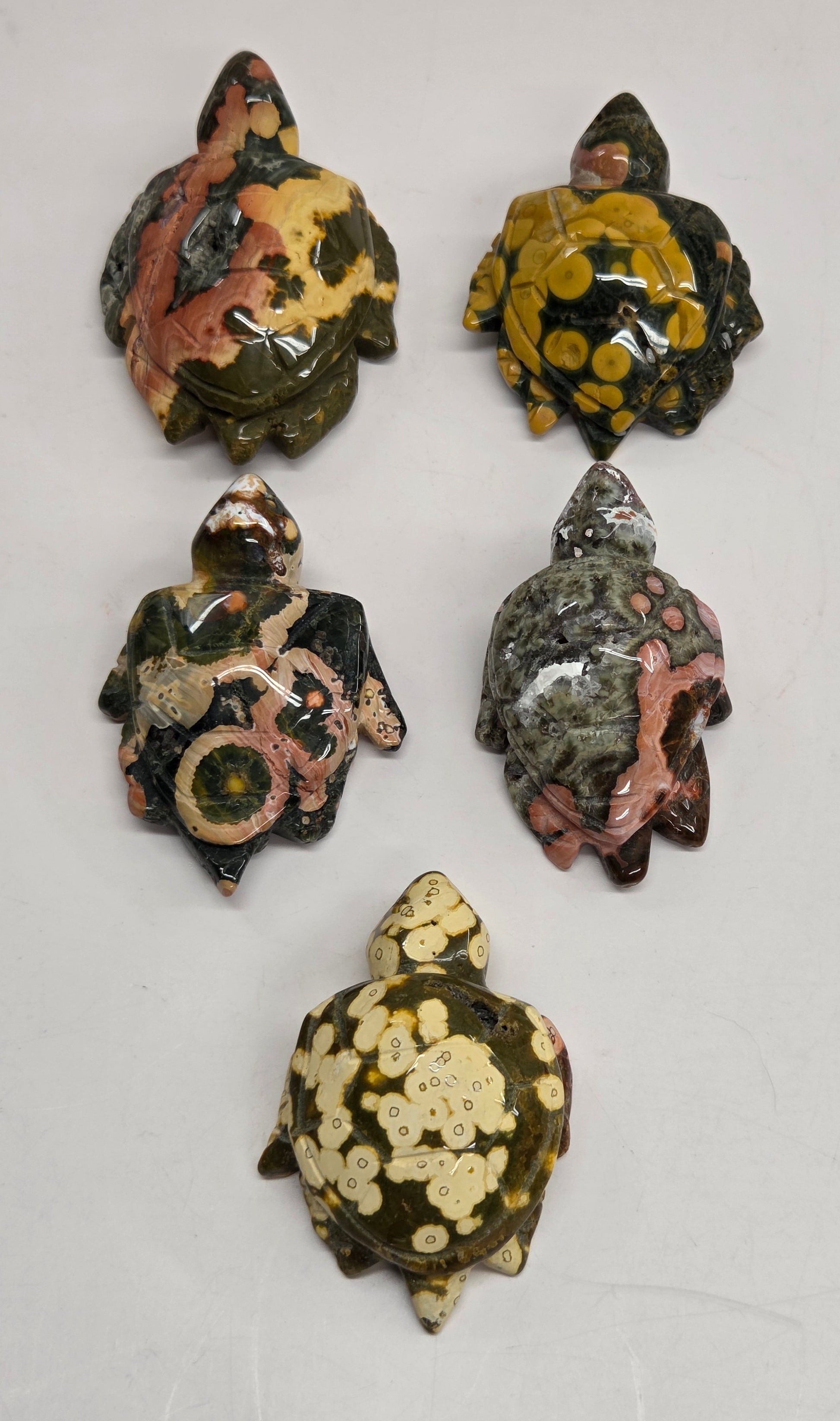 Five small decorative turtle figurines with marbled, mottled patterns in olive green, black, cream, pink, and brown, arranged on a plain light gray surface.