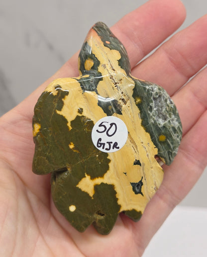 A hand holding a polished stone or mineral specimen with a mottled pattern of olive green, dark brown, and pale yellow, featuring a small white sticker with handwritten numbers.