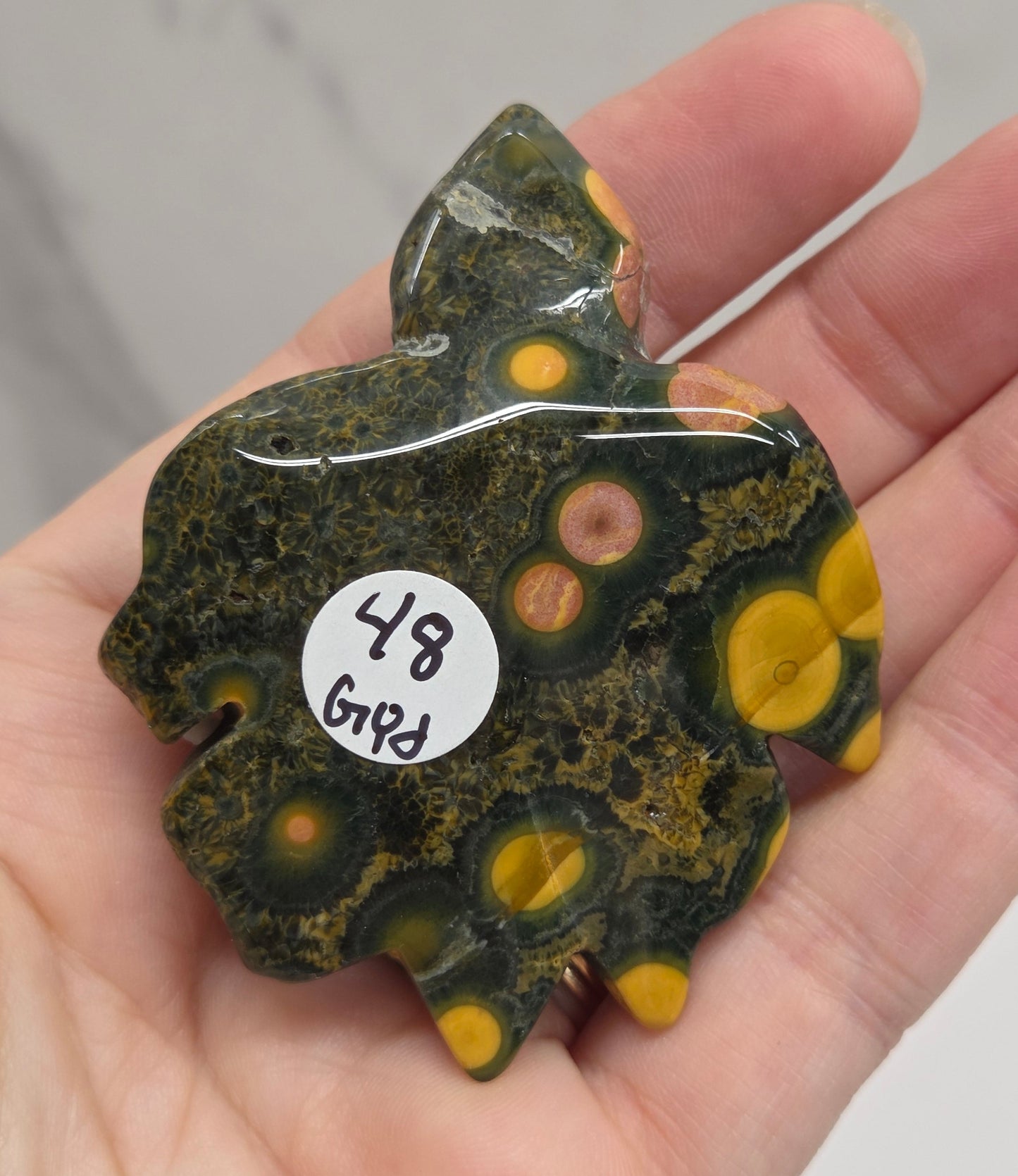 A hand holding a polished natural stone pendant shaped like a turtle, featuring a marbled pattern of dark green, black, and bright yellow-orange spots, with a small white sticker labeled '4.9 GTR' on the side.