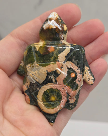 A hand holding a small decorative turtle figurine made from polished stone or glass, showing a marbled pattern of green, beige, and brown.