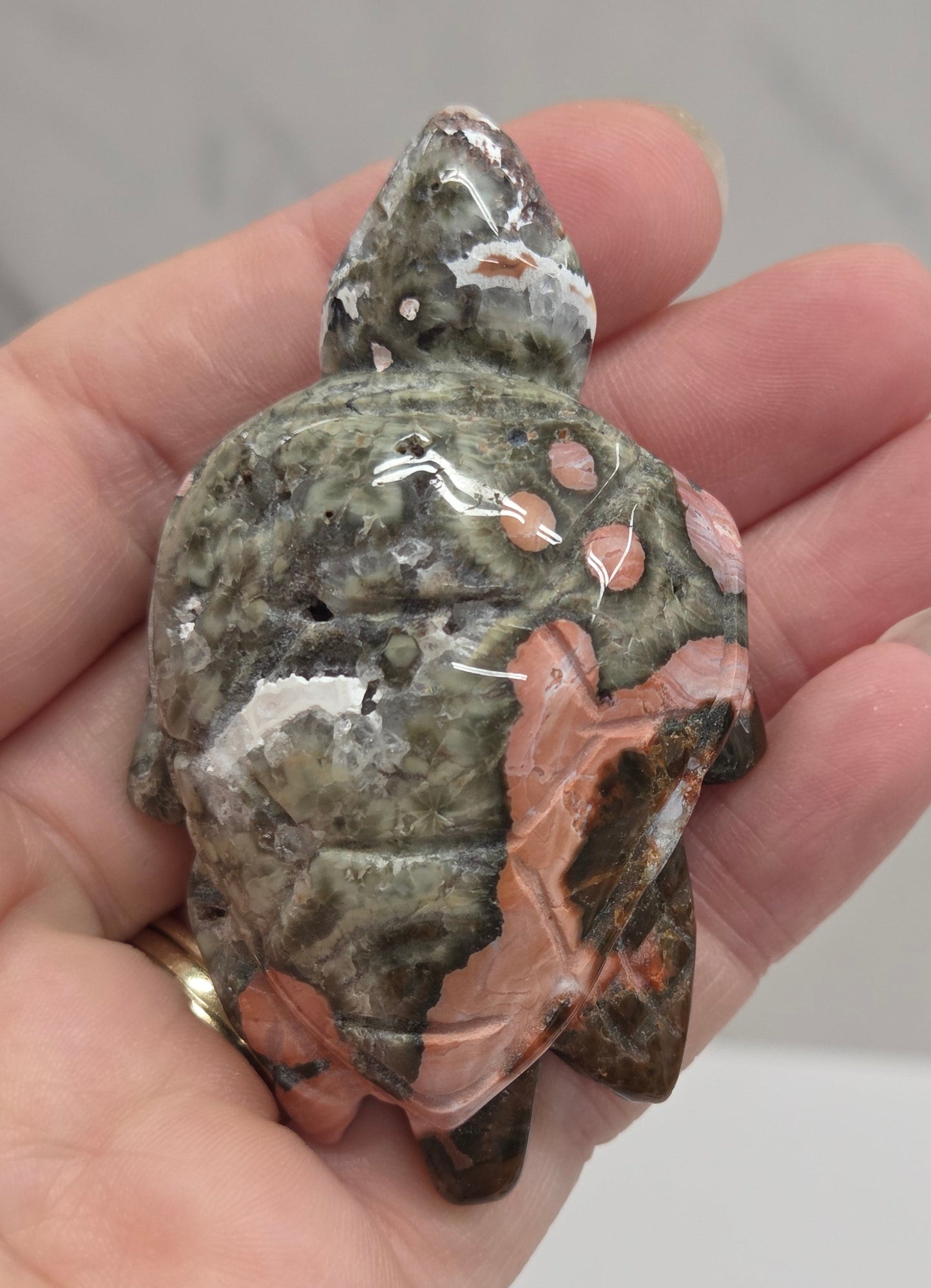 A hand holding a polished carved stone figurine shaped like a turtle, featuring a mottled pattern of gray, green, and peach-pink.