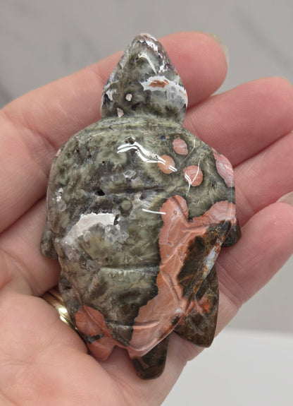 A hand holding a polished carved stone figurine shaped like a turtle, featuring a mottled pattern of gray, green, and peach-pink.