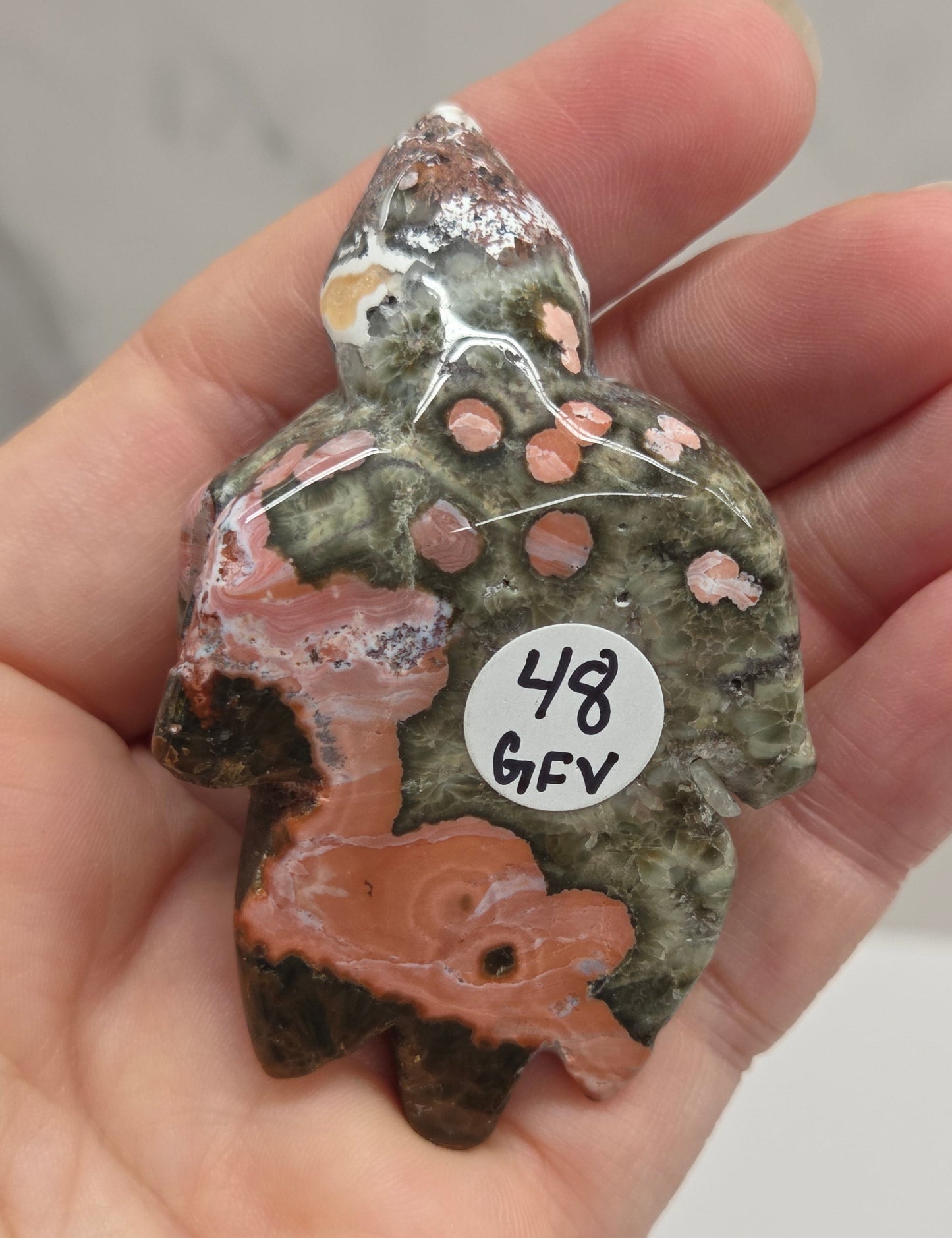 A hand holding a polished natural stone figurine shaped like a small animal, with a white circular price tag reading '48 GGY' attached to it. The stone has mottled green, brown, peach, and black patterns.