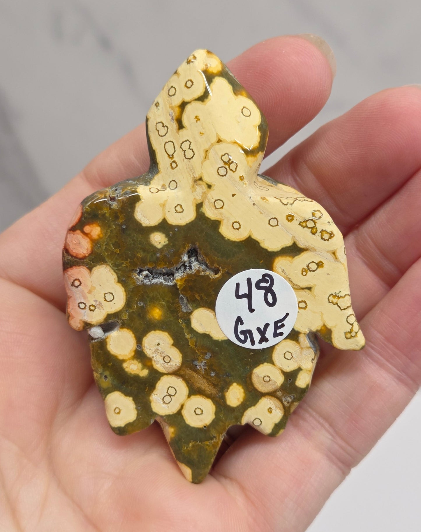 A hand holding a small, irregularly shaped piece of patterned stone or ceramic with a mottled design of olive green, cream, and orange, featuring a small white sticker that reads '13 GTE'.