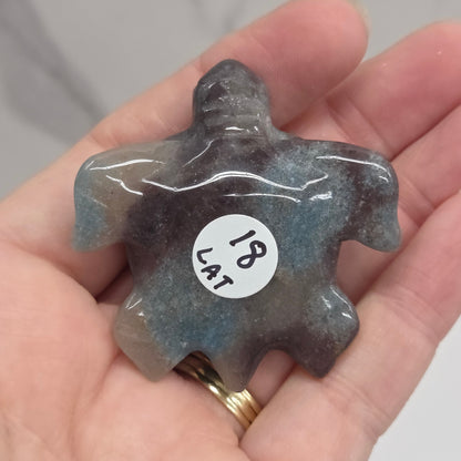 A hand holding a small carved stone figurine shaped like a turtle or sea creature. The stone is dark gray with blue-green iridescent variation.