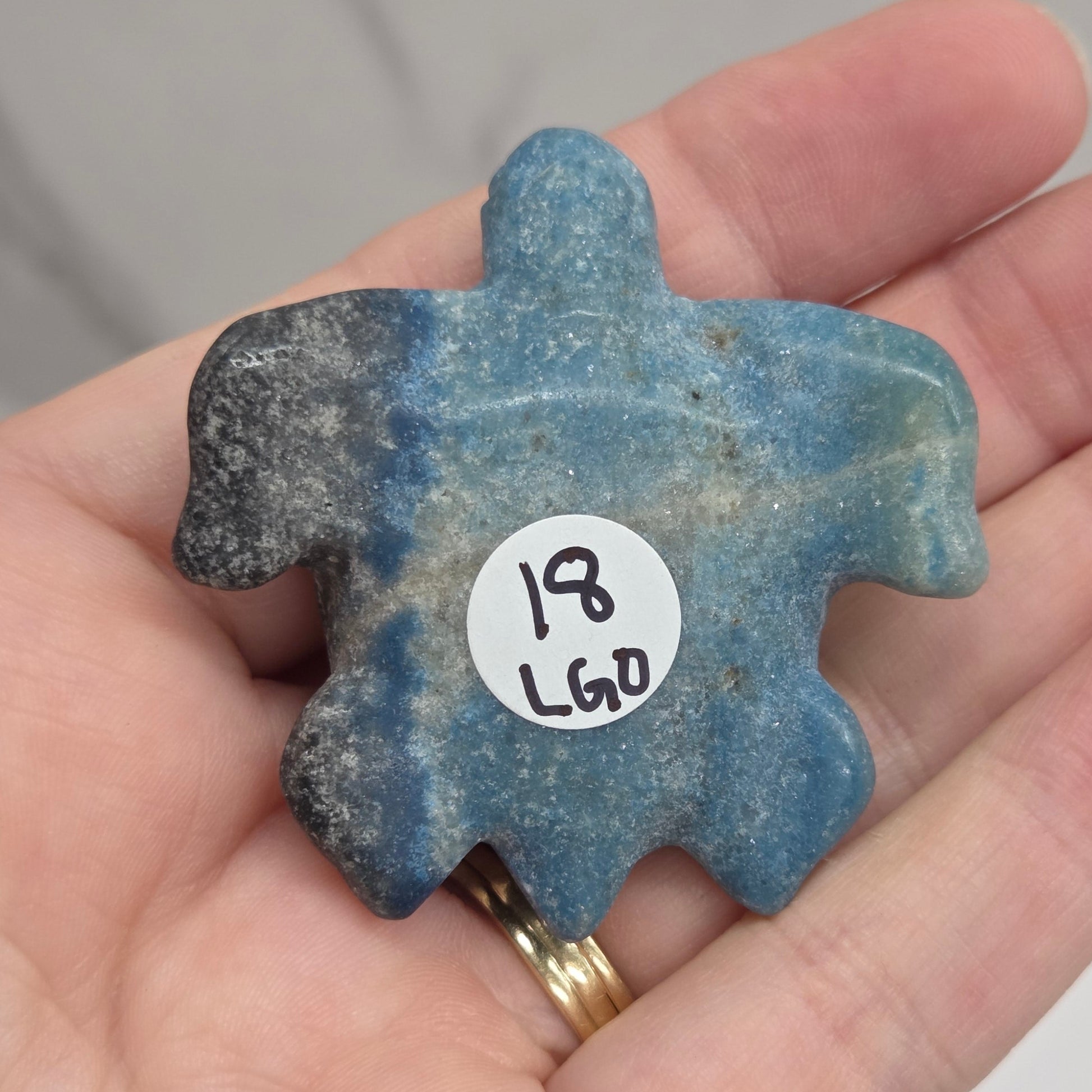 A hand holding a carved blue stone turtle-shaped piece with a small white sticker reading '18 LGO' on the front, against a light marble-like background.
