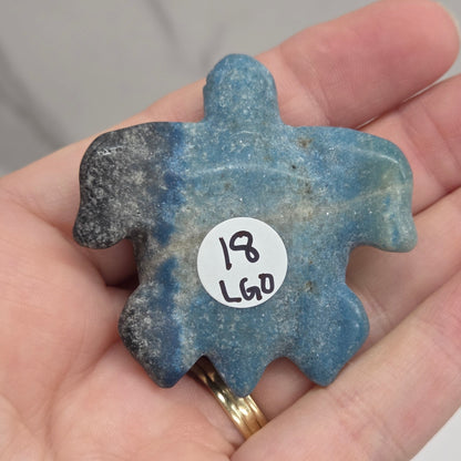 A hand holding a carved blue stone turtle-shaped piece with a small white sticker reading '18 LGO' on the front, against a light marble-like background.