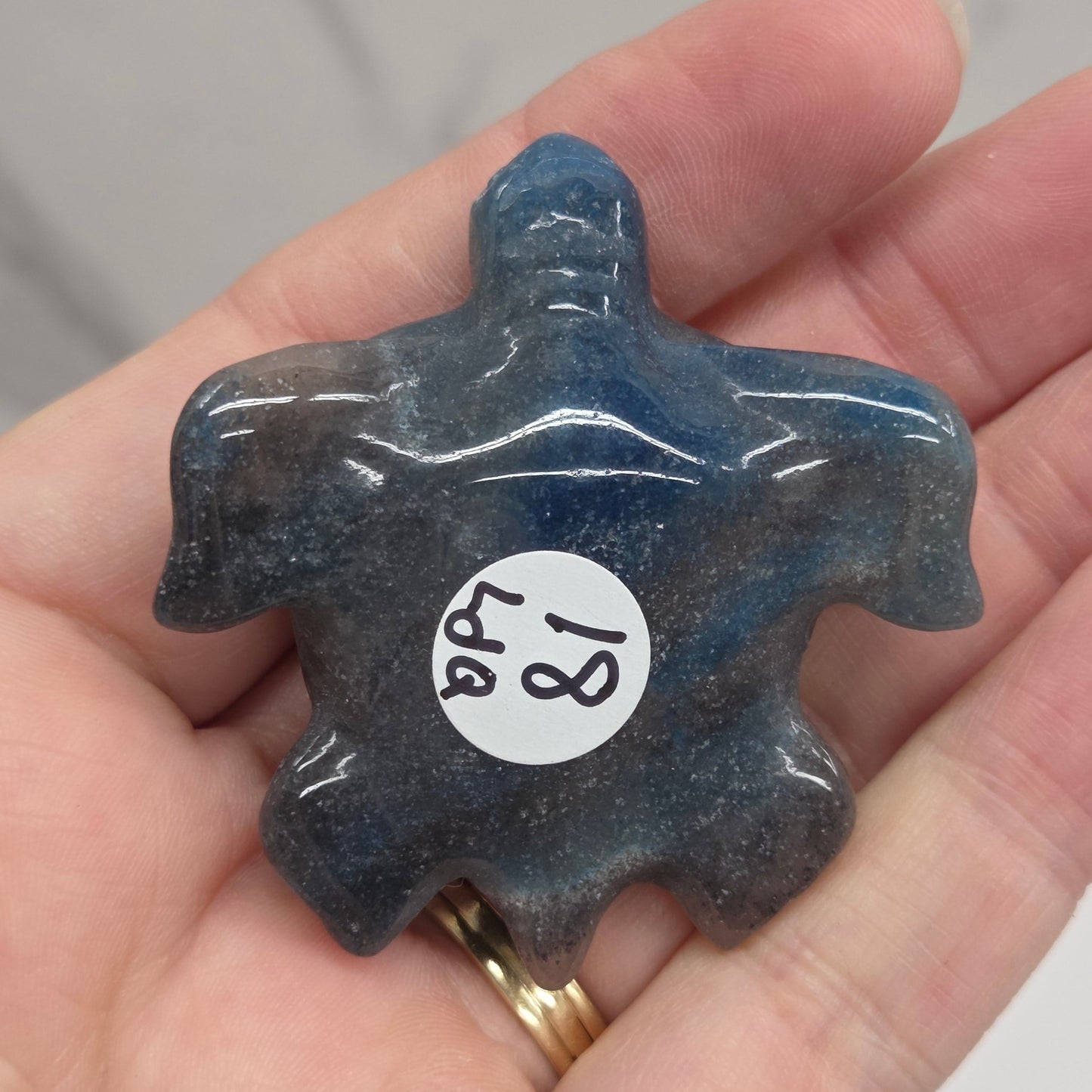 A hand holding a small carved dark blue stone figurine with a white circular sticker marked '18 48' on it, against a light marble-like background.