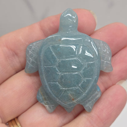 A small carved turtle-shaped pendant or charm in a pale blue-gray stone, held in a person's hand. The hand has a gold ring on the ring finger, and the background is a light gray marble-like surface.