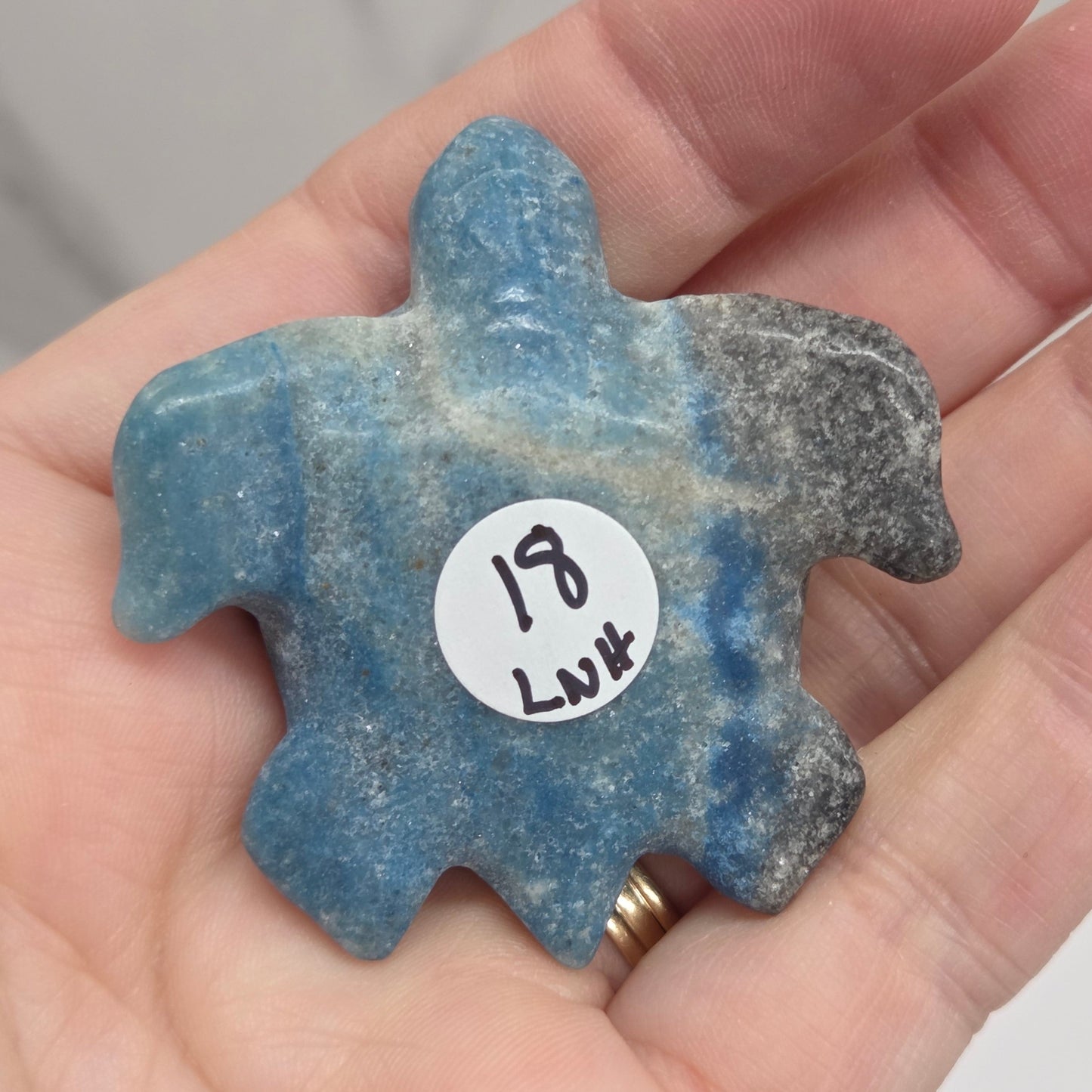 A hand holding a small, irregularly shaped blue stone pendant or charm with a white price sticker on it, against a light gray marble background.