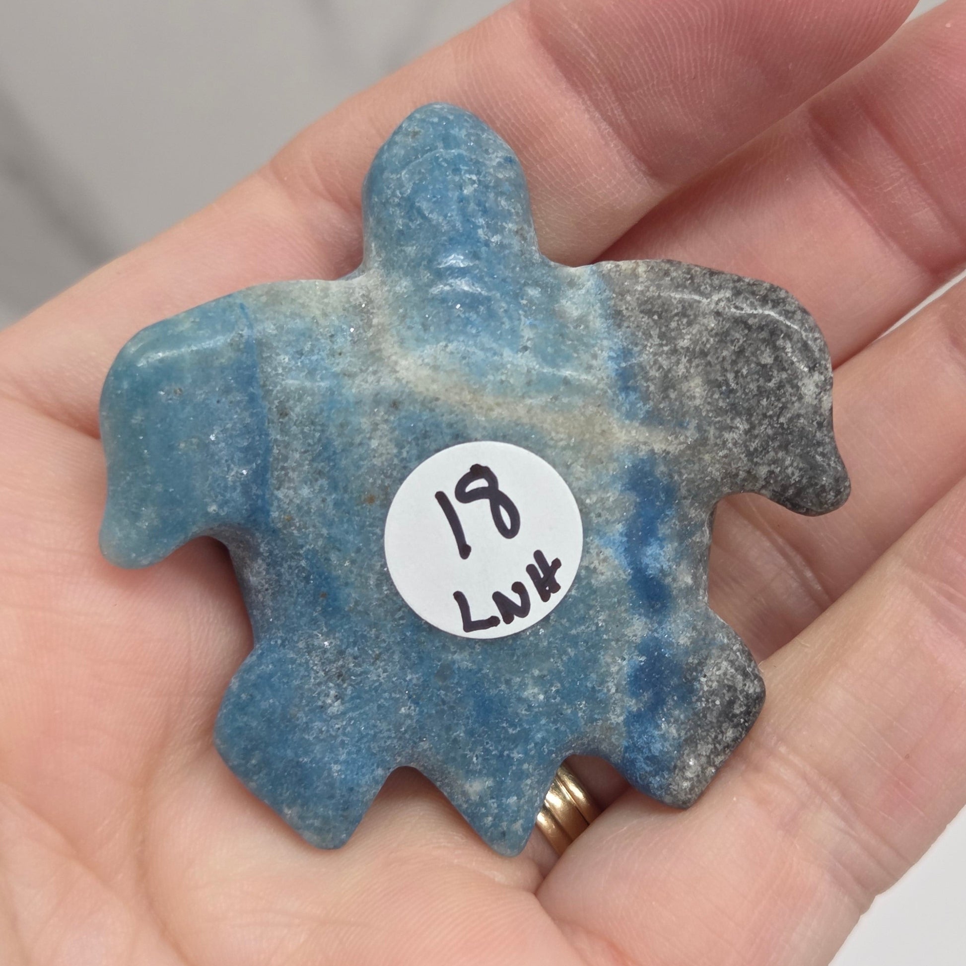 A hand holding a small, irregularly shaped blue stone pendant or charm with a white price sticker on it, against a light gray marble background.