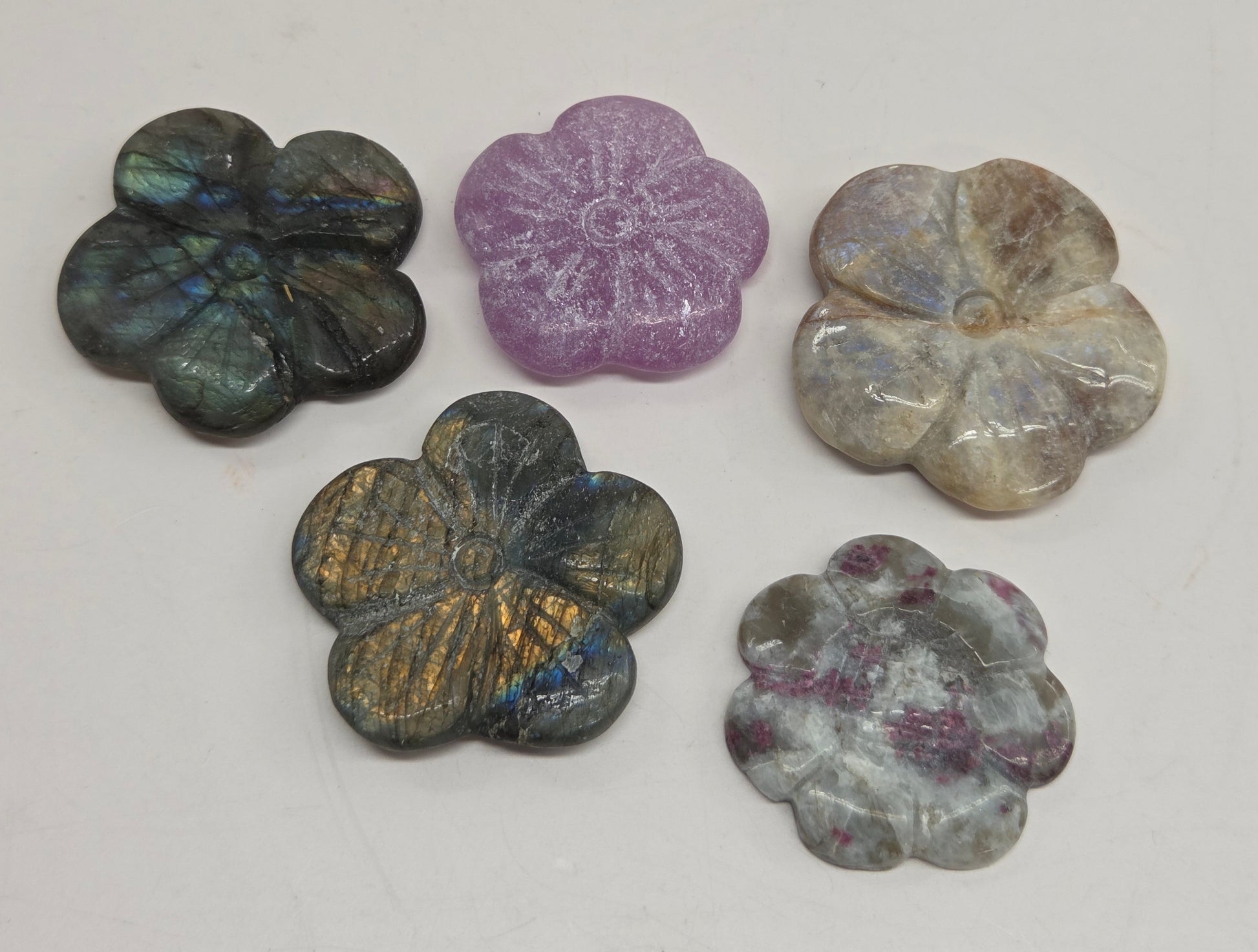 Five carved flower-shaped carvings made from natural stones, including Labrador, amethyst, and rose quartz, arranged on a plain off-white surface.