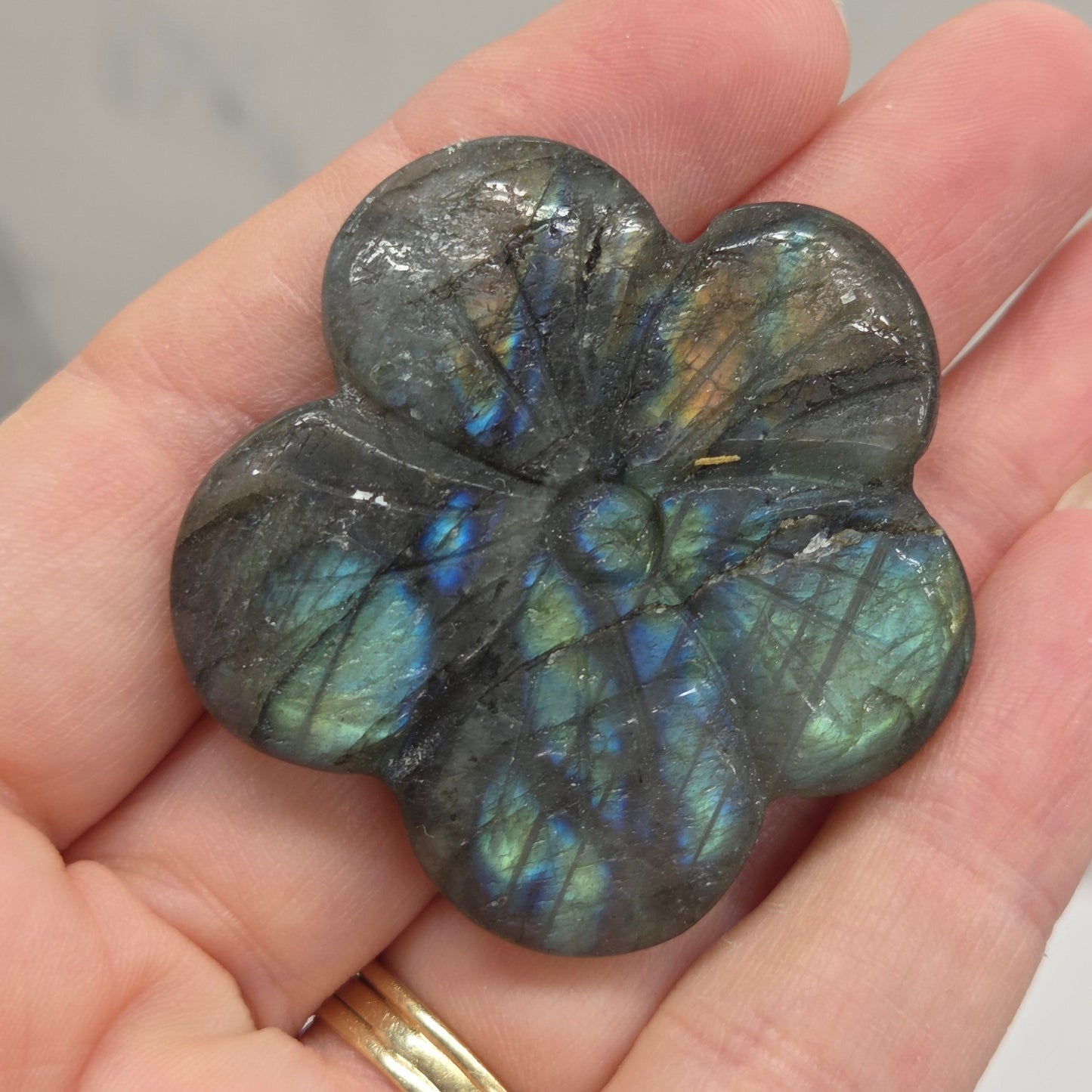 A five-petal flower-shaped labradorite carving held in a person's hand, showing its characteristic green and blue iridescent flash against a light gray marble background.