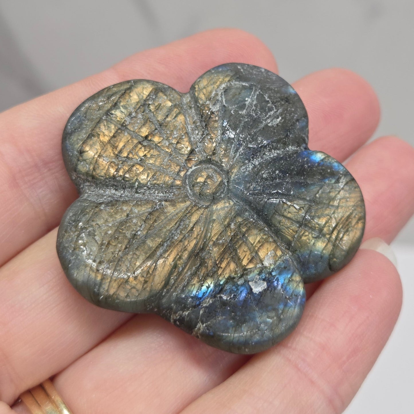 A five-petal flower-shaped labradorite stone charm held in a person's hand. The stone displays a metallic gold and gray crystalline structure with a blue iridescent flash under light.