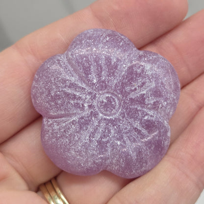 A lavender purple, flower-shaped translucent stone or crystal held in a person's hand. The crystal has a textured, glittery surface and is shown against a soft-focus gray background.
