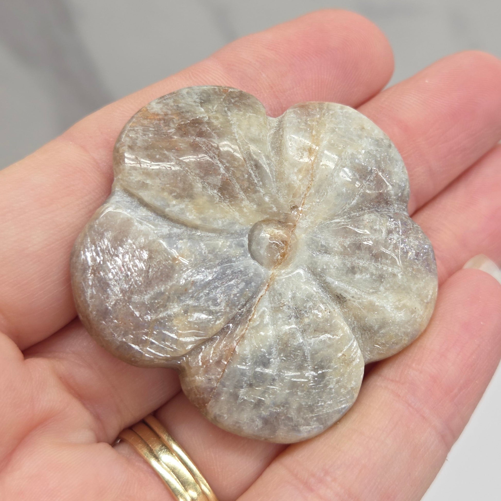 A carved stone flower pendant with a textured, polished surface, held in a person's hand. The pendant has a natural stone appearance with beige, gray, and brown tones.