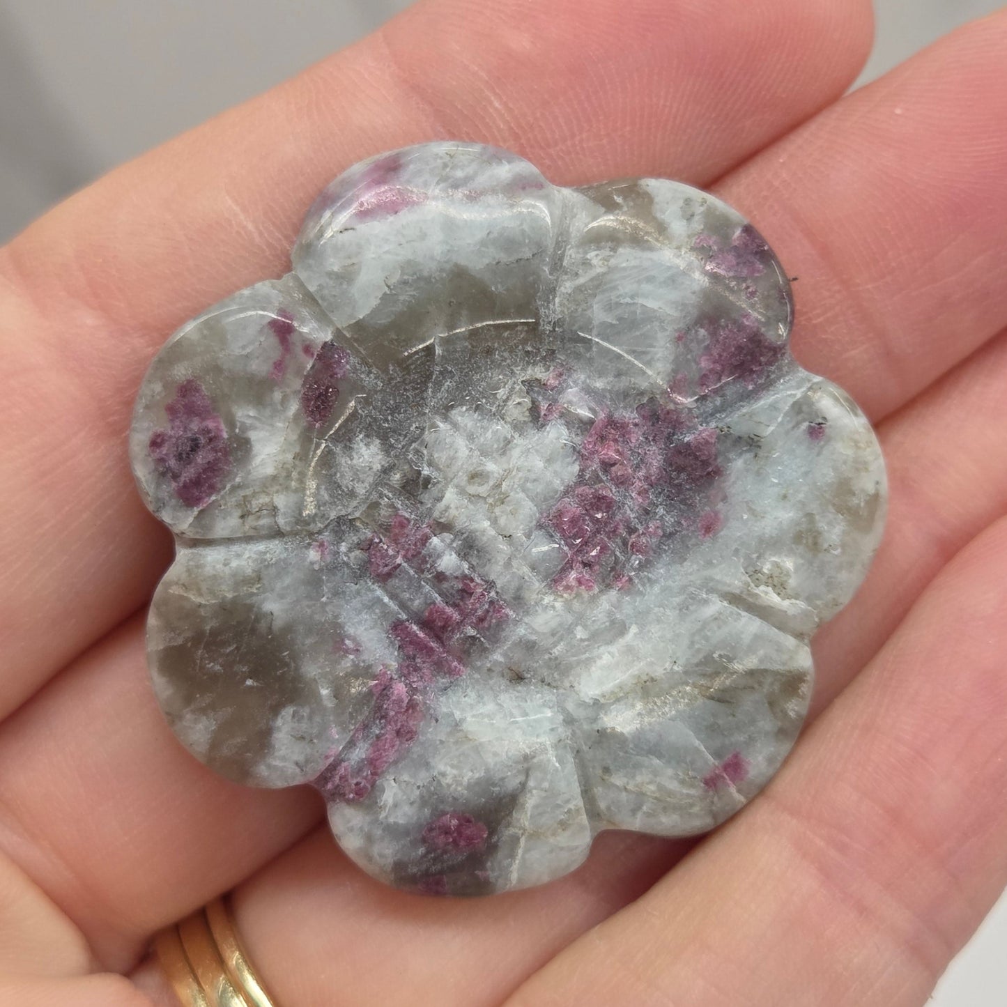 A hand holding a carved flower-shaped gemstone with a mottled pattern of gray, white, and purple crystals, with a gold ring on the finger.