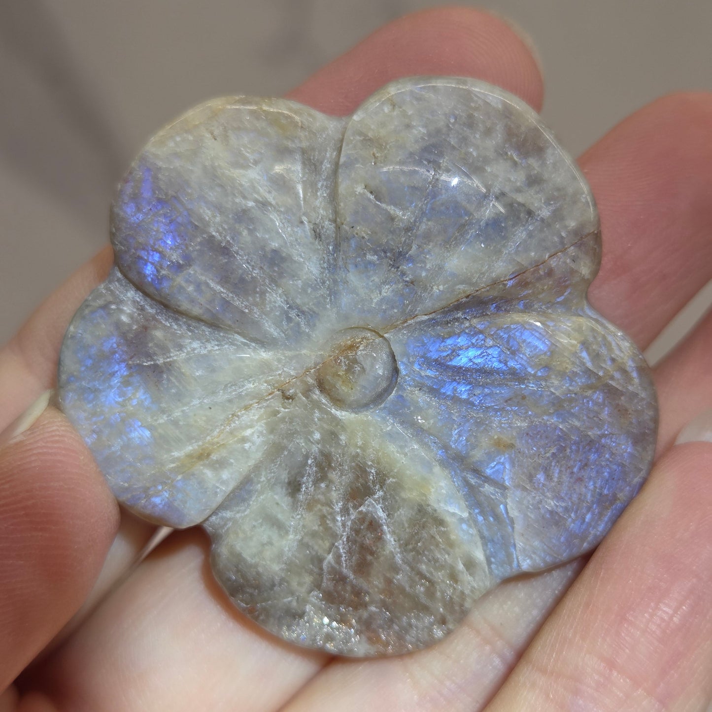 A carved stone flower-shaped pendant or charm with a five-petal design, held in a person's hand. The stone exhibits a grayish-brown base color with a strong blue iridescent play-of-color effect near the center and lower petals.