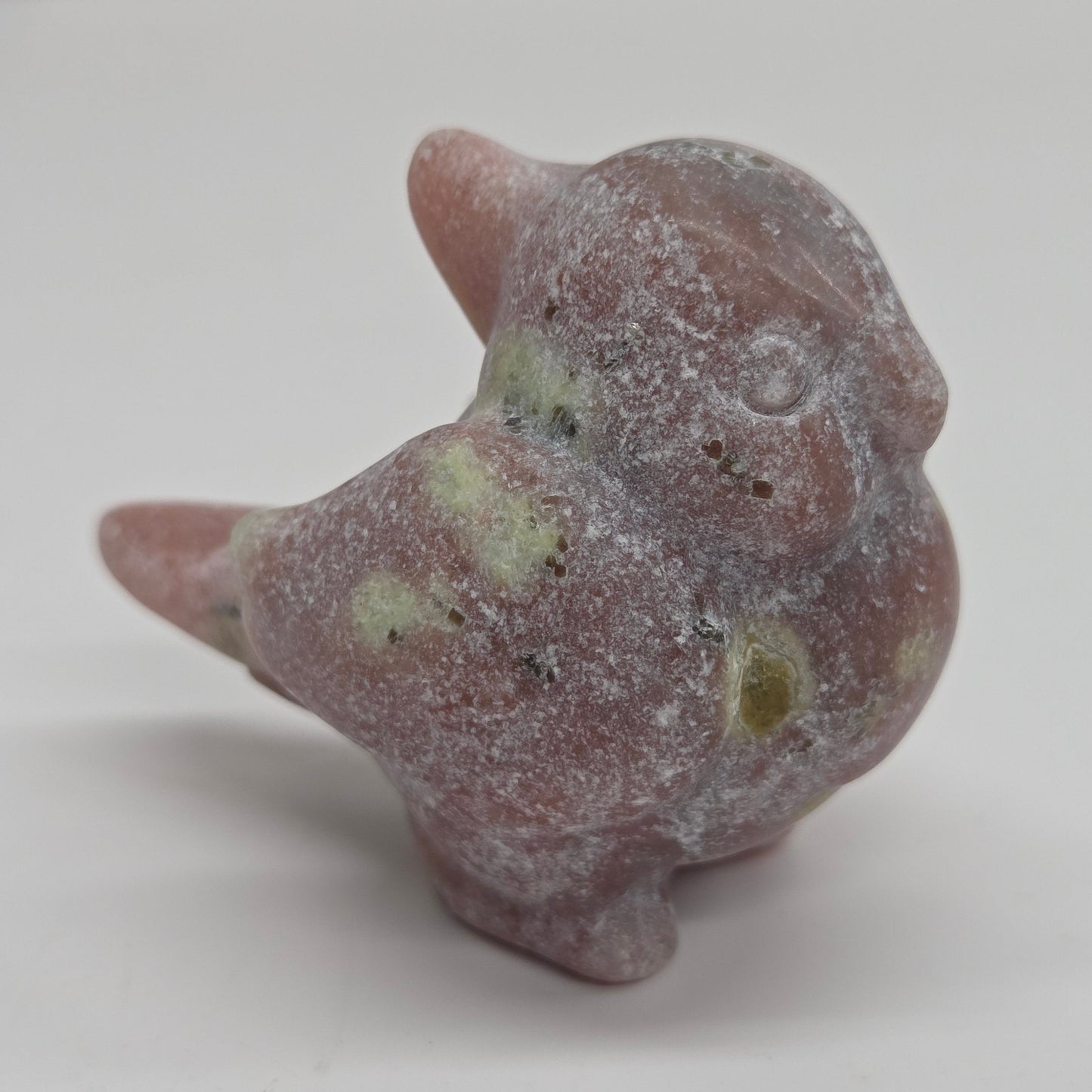 A small carved figurine of a bird, likely made of stone or resin, with a dusty pinkish-brown color and light green marbled patches, set against a plain light gray background.