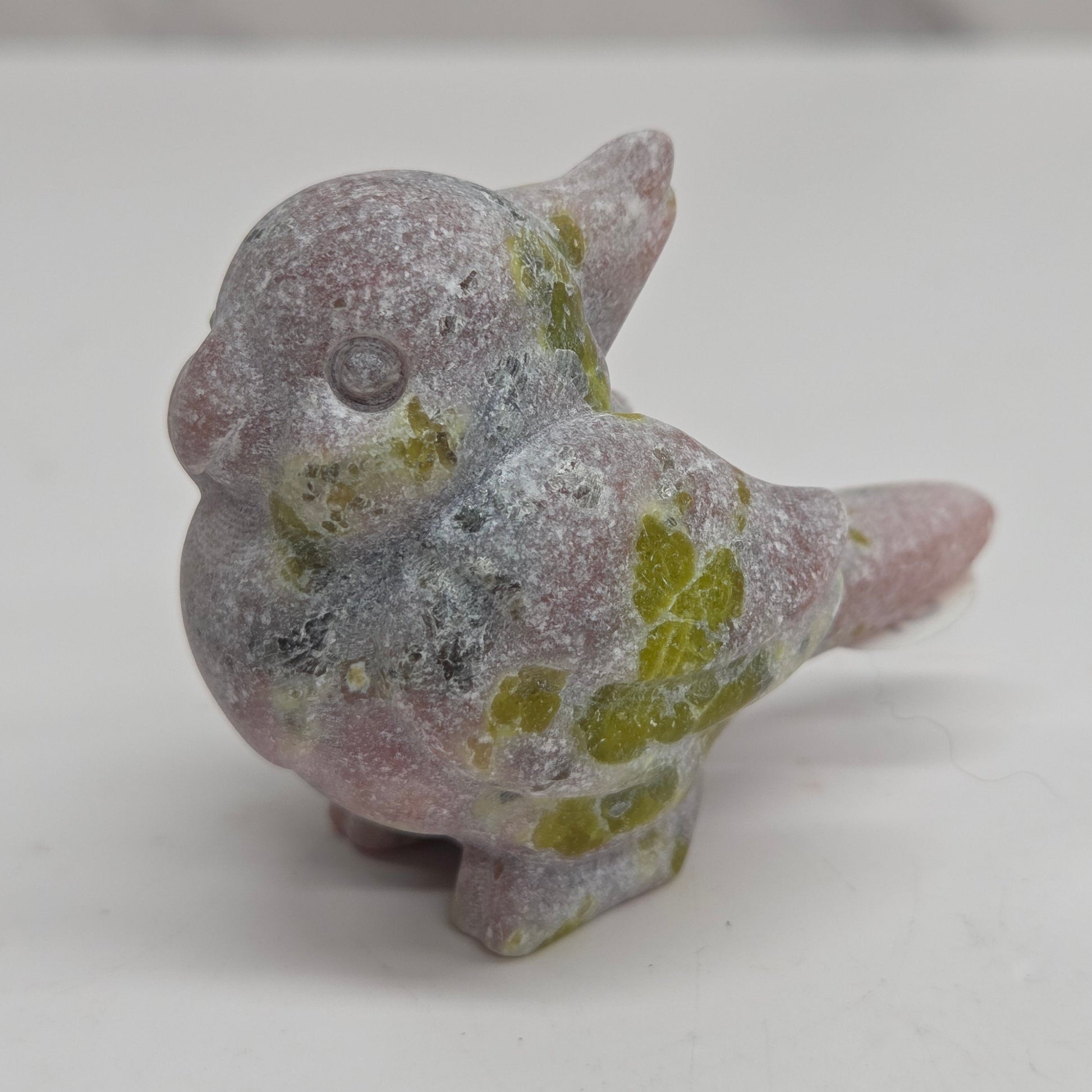A small bird figurine made of textured stone, featuring a dusty lavender-pink body with patches of olive green and gray, standing on a white surface.