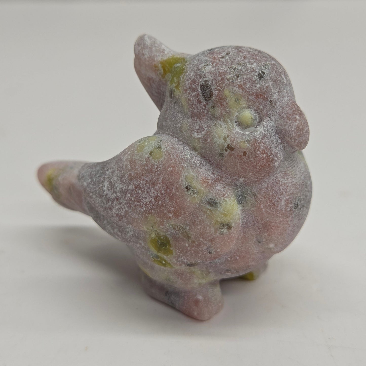 A small carved bird figurine made of pinkish stone with green and yellowish inclusions, standing on a plain light surface.