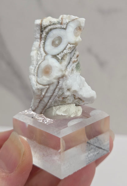 A hand holding a clear faceted crystal cube attached to a white and gray agate stone piece, shown against a light marble-patterned background.