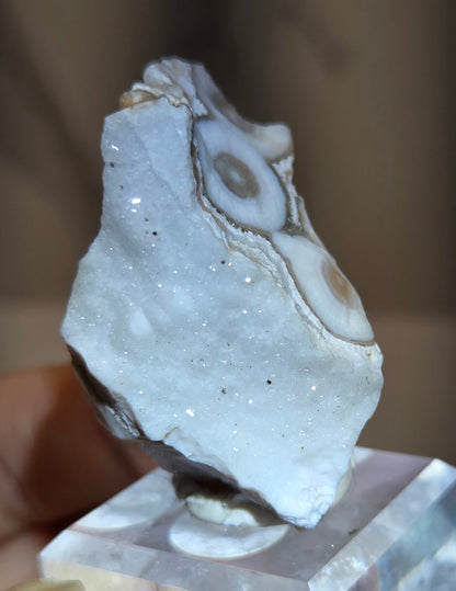 A hand holding a raw agate crystal specimen with distinctive banded eye-like patterns, next to a clear quartz crystal cube against a blurred brown background.