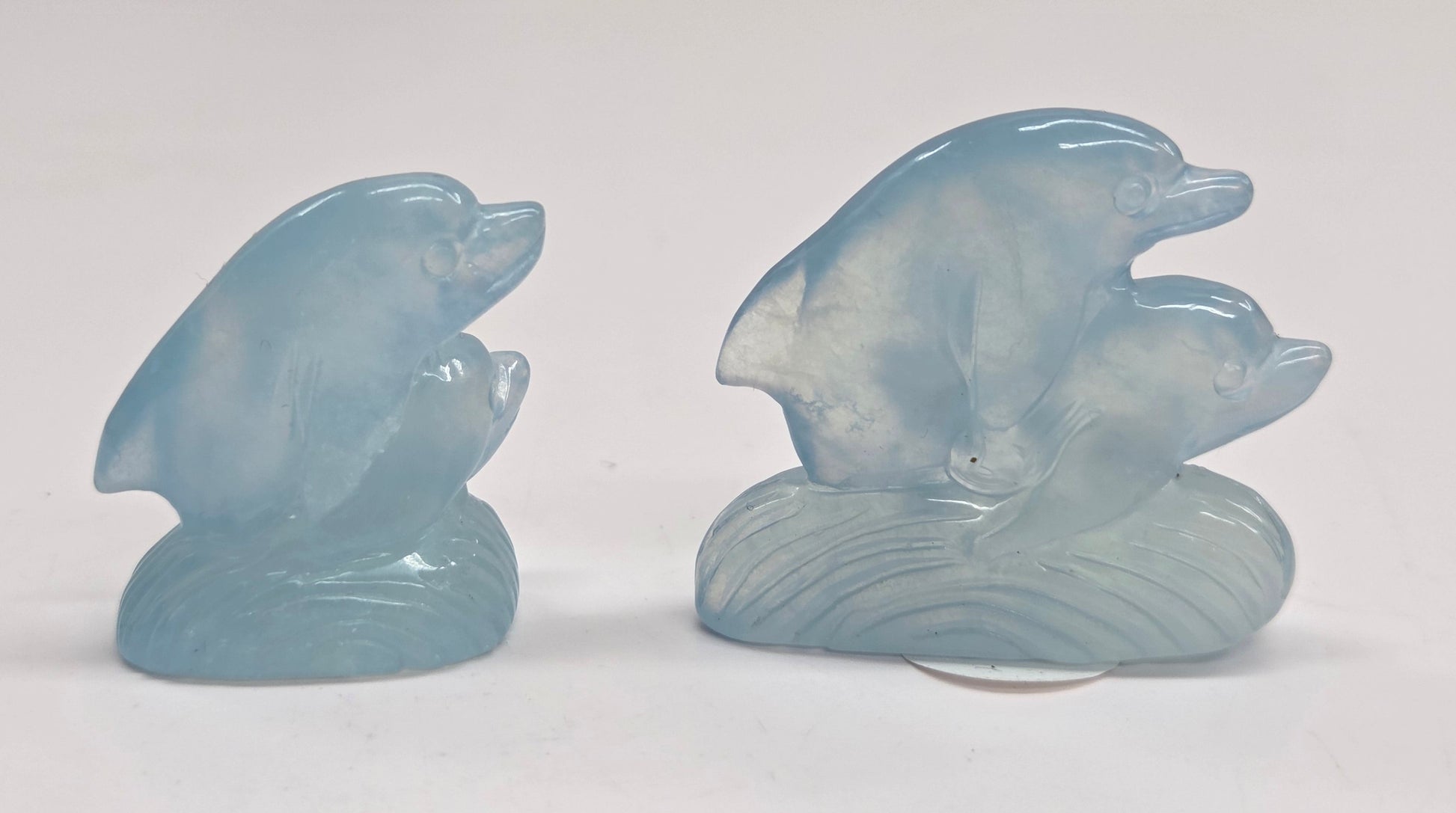 Two light blue carved stone figurines, two dolphins, posed together on a matching wave-shaped base against a plain white background.
