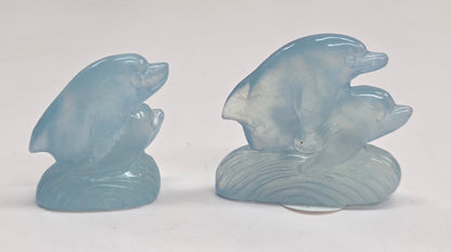 Two light blue carved stone figurines, two dolphins, posed together on a matching wave-shaped base against a plain white background.