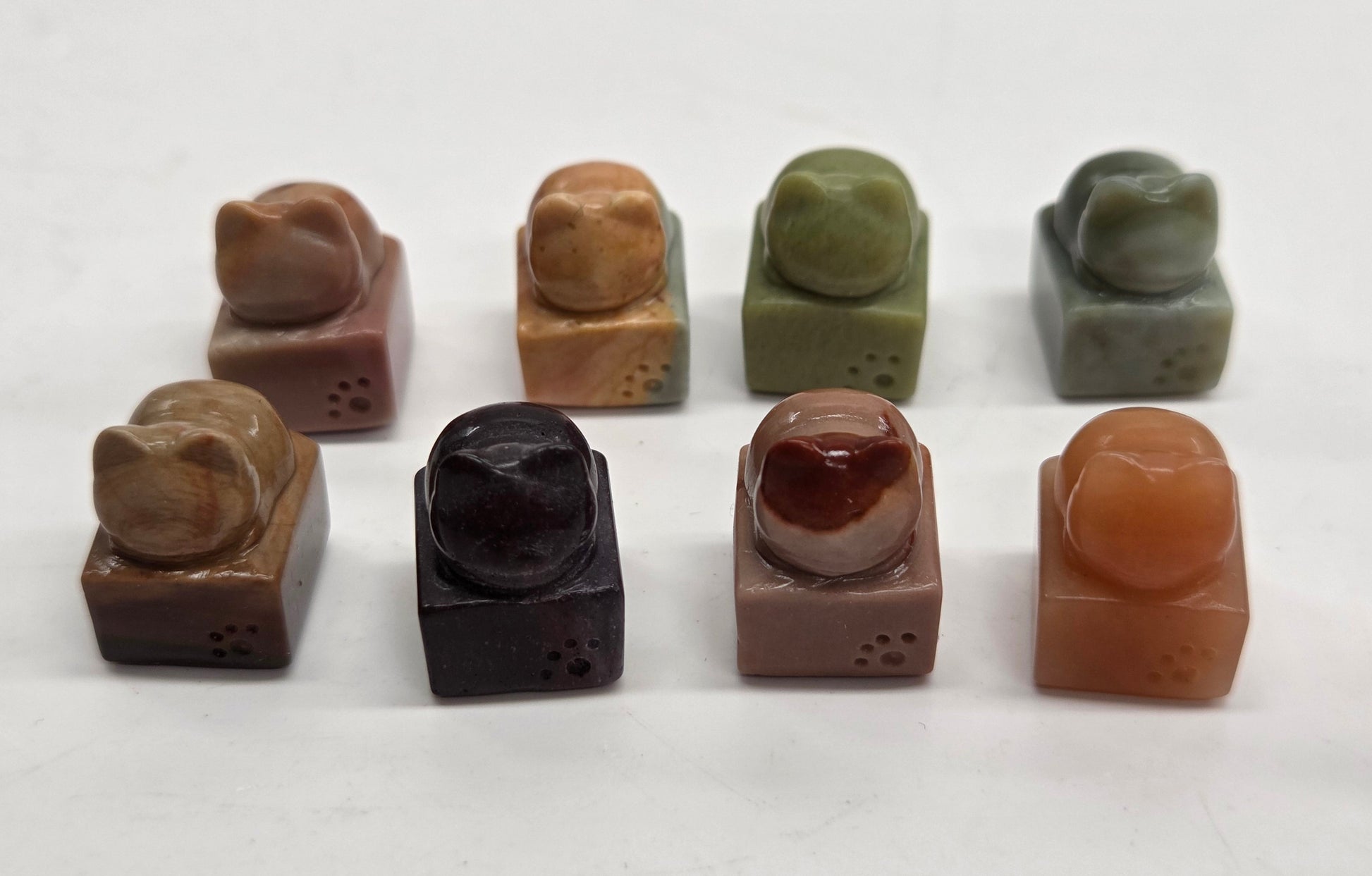Eight small carved stone figurines arranged in two rows on a plain white background. The figures appear to be stylized animal or creature motifs in various earthy stone colors including brown, tan, olive green, charcoal black, and terracotta.