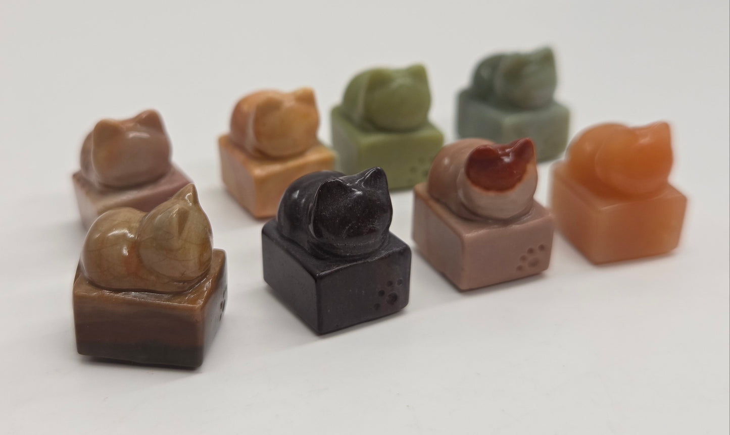 A set of small carved stone figurines shaped like sleeping cats, displayed on a plain white background. The set includes stones in brown, black, green, orange, and beige.