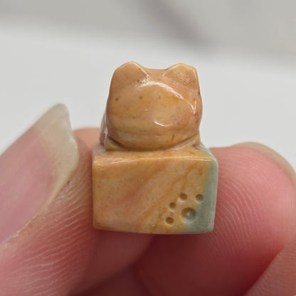 A small carved stone charm or bead, shaped like a cat beside a square piece with three holes, held between a person's fingers against a light gray background.