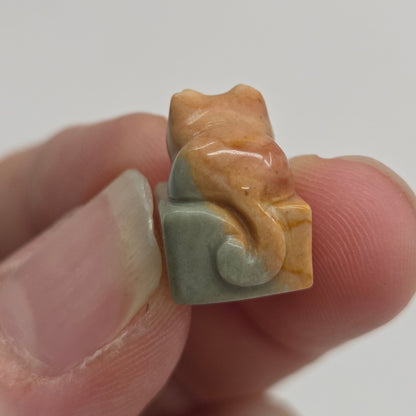 A hand holding a small carved figurine of a cat sitting on a block, made from a marbled stone with orange, yellow, and pale green tones.