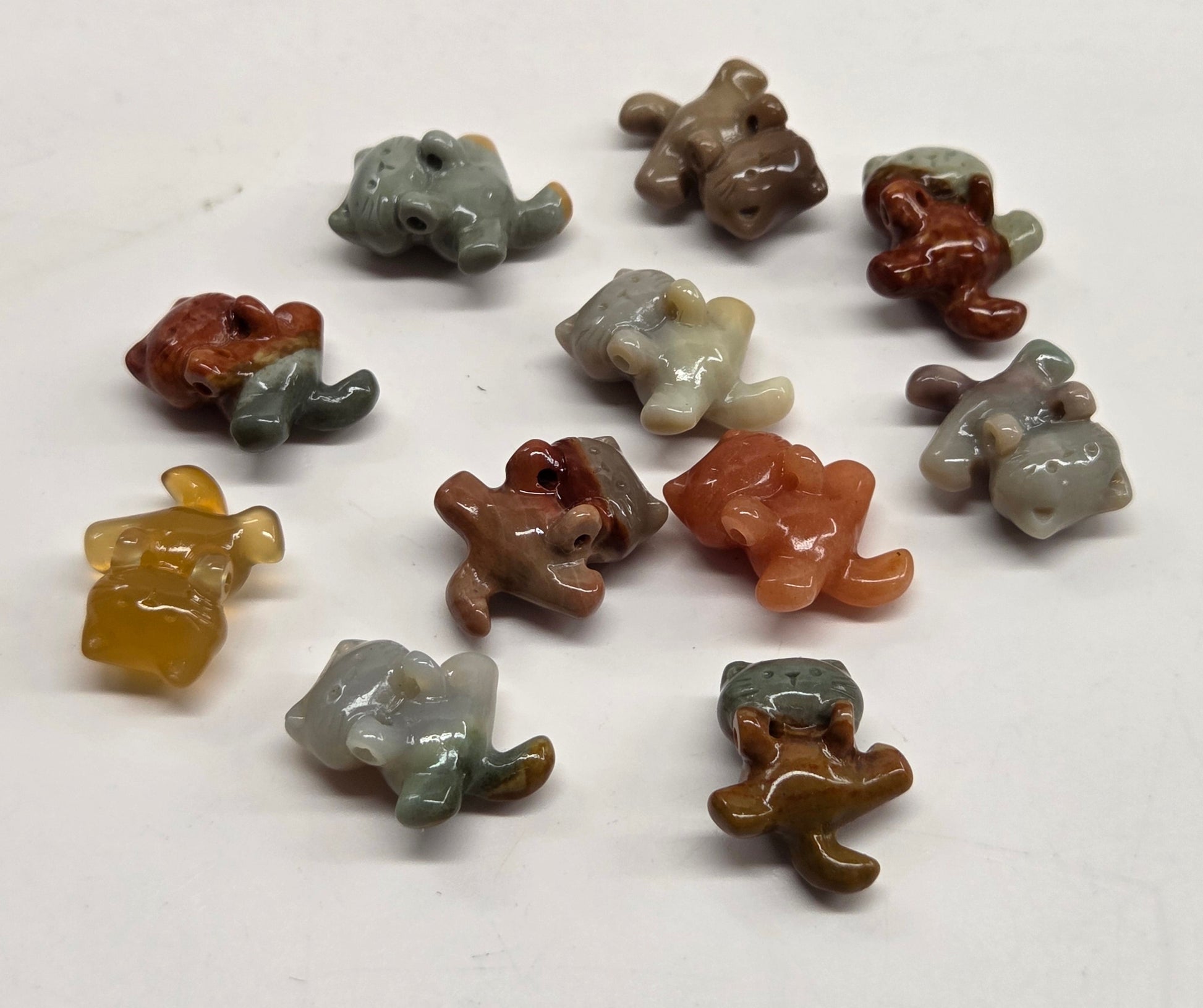 A group of small, hand-carved cat beads made from agate or jasper in various earthy colors like brown, gray, orange, and yellow, arranged on a plain white surface.