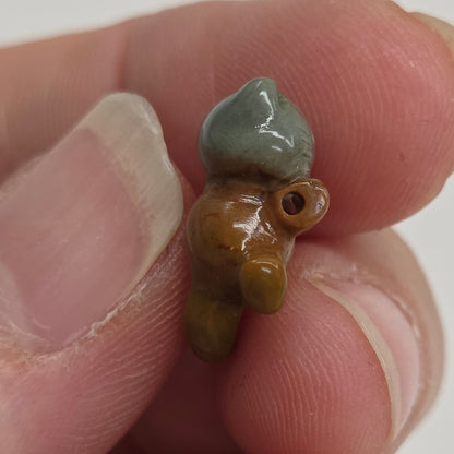 A tiny handcrafted bead or charm being held between fingers. The bead is brown with a small mint green accent on one end, photographed close-up against a plain light background.