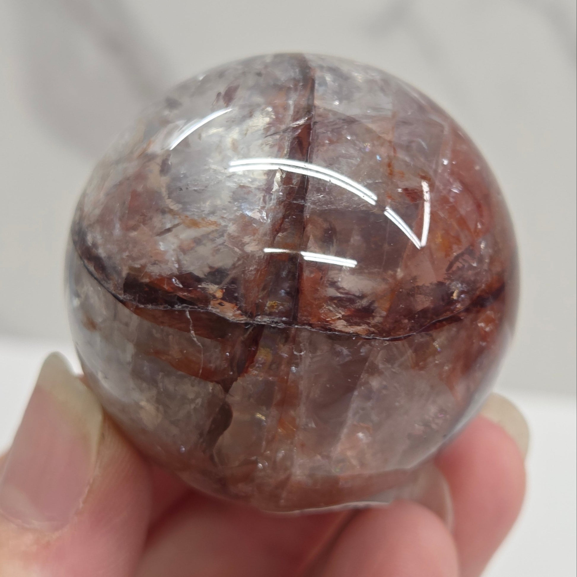 A hand holding a polished round crystal sphere with brown and gray translucent stone showing natural fissures and inclusions.