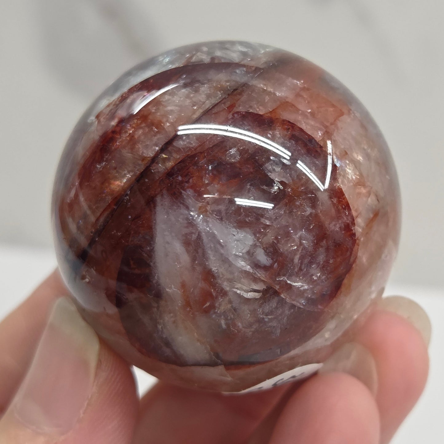A hand holding a polished round crystal sphere with swirling brown, reddish-brown, and translucent white patterns, photographed against a light marble-like background.
