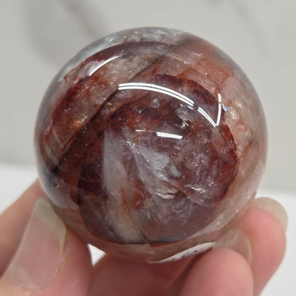 A hand holding a polished round crystal sphere with swirling brown, reddish-brown, and translucent white patterns, photographed against a light marble-like background.