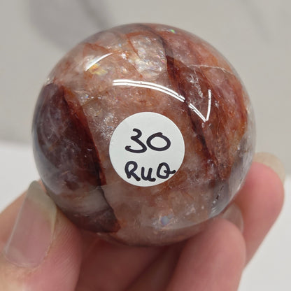 A hand holding a polished round stone with a white sticker that reads '30 RUG' on a light marble-like surface.