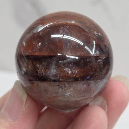 A polished brown gemstone sphere held by a hand against a light gray marble background.