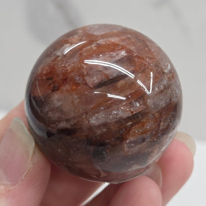A hand holding a polished round brown stone with natural marbled patterns, photographed close-up against a light marble-like background.