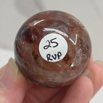 A hand holding a polished brown stone sphere with a white circular sticker that reads '25 002'.