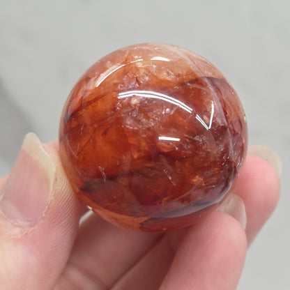 A hand holding a polished round orange-brown stone or crystal sphere against a light gray marble-like background.