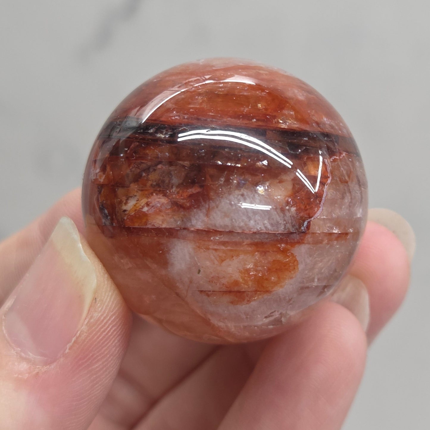 A hand holding a polished round gemstone with a warm reddish-brown and orange coloration and white cloudy inclusions, set against a light gray marble-patterned background.