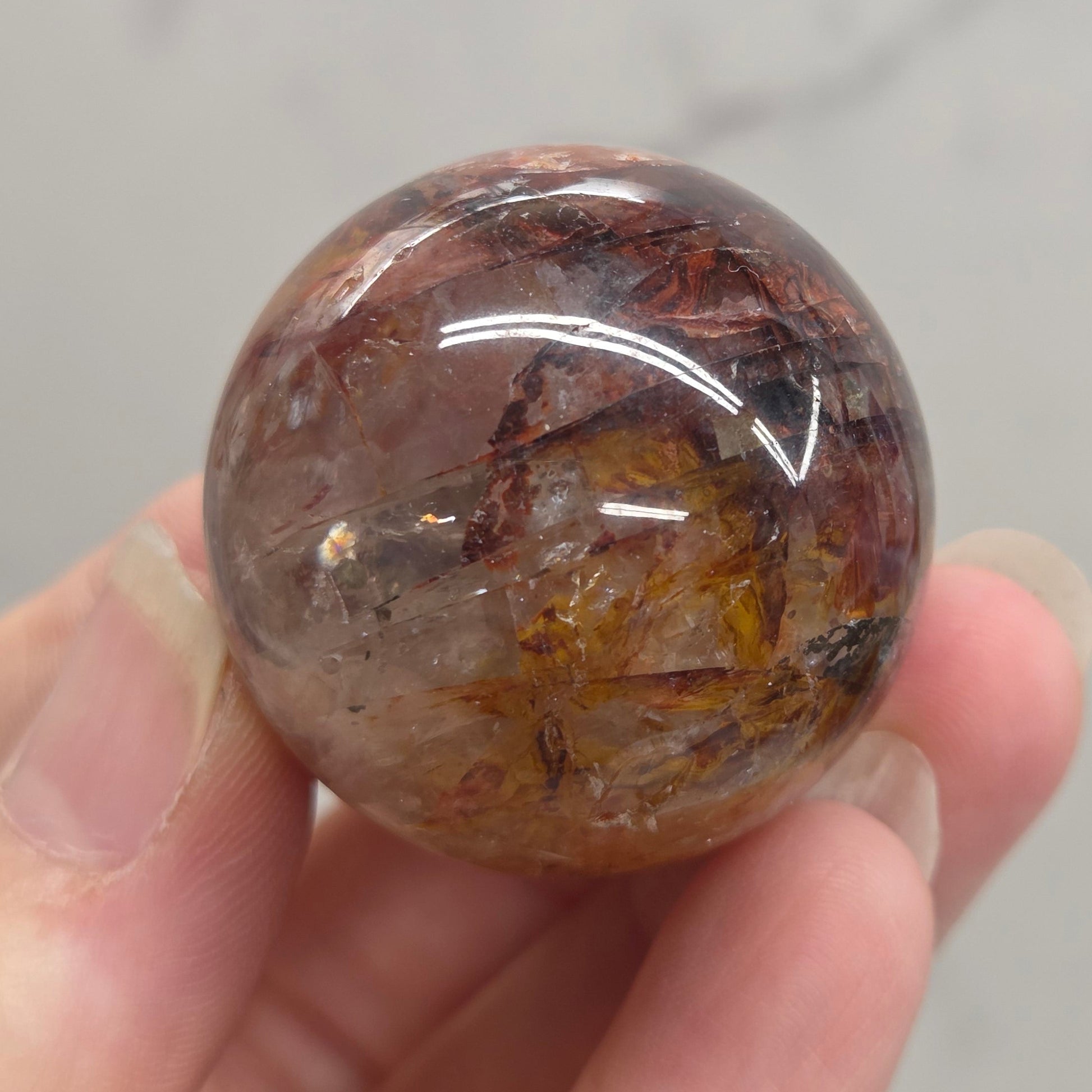 A hand holding a polished round crystal sphere with internal inclusions, shown against a light gray marble-like background.