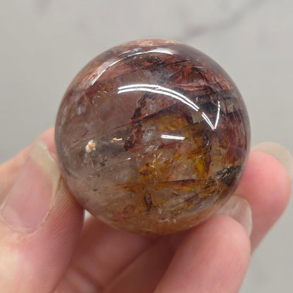 A hand holding a polished round crystal sphere with internal inclusions, shown against a light gray marble-like background.