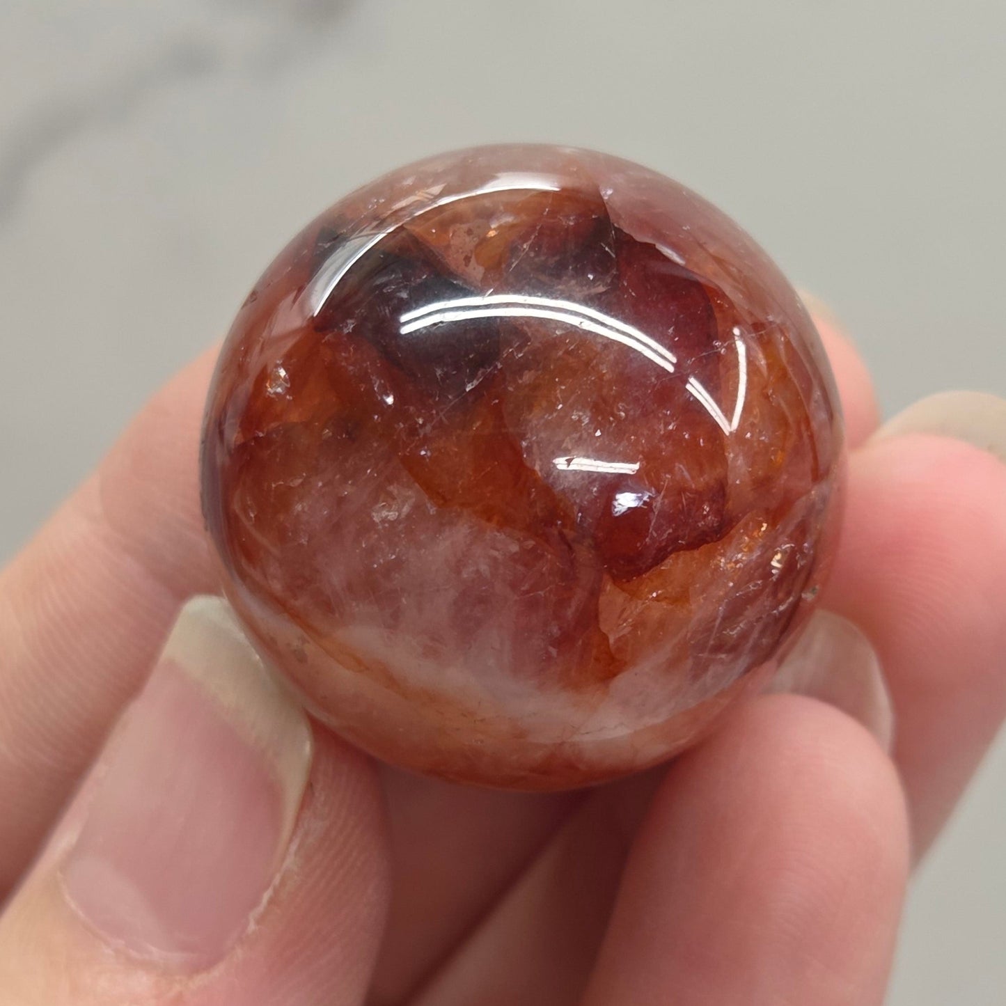 A hand holding a polished round gemstone with a warm reddish-brown and amber coloration, set against a light gray marble-patterned background.
