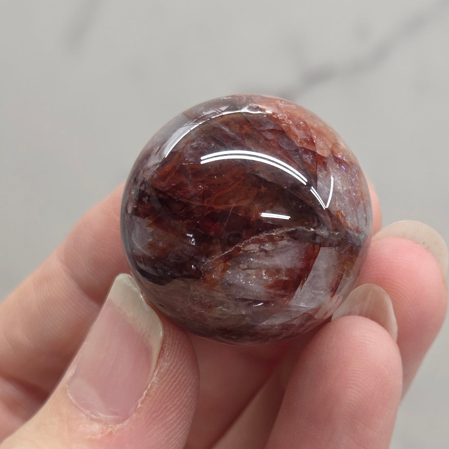 A hand holding a polished round gemstone with a marbled pattern of reddish-brown, dark brown, and black, set against a light gray marble background.