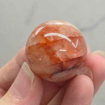 A hand holding a polished round gemstone with warm orange, peach, and brown marbled patterns against a light gray marble background.